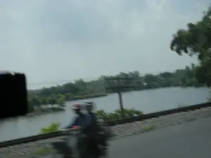 Bức ảnh bị mờ do được chụp từ một phương tiện đang di chuyển, ghi lại một khung cảnh nông thôn. Ở phía trước, hai người đội mũ bảo hiểm đang đi xe máy trên một con đường nhựa. Song song với con đường là một đoạn đường ray xe lửa. Phía sau họ là một con sông (hoặc hồ) rộng, bên kia bờ là cây cối xanh tươi. Một cột điện cũng có thể được nhìn thấy gần bờ sông. Bầu trời u ám và có nhiều mây.。（日本語訳）。写真は移動中の乗り物から撮影されたためぼやけており、田舎の風景を捉えています。手前では、ヘルメットをかぶった2人がアスファルトの道路をバイクで走っています。道路と平行して線路が走っています。彼らの後ろには広い川（または湖）があり、対岸には青々とした木々があります。川岸の近くには電柱も見えます。空はどんよりとしており、雲が多いです。