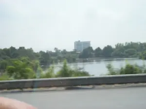 Bức ảnh chụp phong cảnh nhìn từ một phương tiện đang di chuyển, có thể là ô tô hoặc xe buýt. Ở phía trước là một phần của con đường và lan can bảo vệ bằng kim loại. Phía sau lan can là một hồ nước hoặc một khúc sông rộng, được bao quanh bởi cây cối xanh tươi. Ở phía xa, có một tòa nhà bê tông lớn, màu trắng, nhiều tầng. Bầu trời có màu trắng đục và toàn bộ khung cảnh hơi mờ, cho thấy ảnh được chụp qua cửa sổ khi xe đang chạy.。（日本語訳）。移動中の乗り物、おそらく車かバスから見た風景を撮影した写真です。前方には道路の一部と金属製のガードレールがあります。ガードレールの後ろには、青々とした木々に囲まれた湖または広大な川の一部があります。遠くには、白くて大きな多層階のコンクリートの建物があります。空は乳白色で、風景全体が少しぼやけており、車が走行中に窓越しに撮影されたことを示しています。