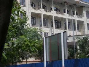 Bức ảnh chụp một tòa nhà lớn, nhiều tầng, có kiến trúc bề thế, mang phong cách cổ điển hoặc thuộc địa. Tòa nhà có màu kem nhạt, nổi bật với hàng cột tròn lớn chống đỡ các tầng trên và ban công. Có rất nhiều cửa sổ bằng kính với khung màu sẫm. Phía trước tòa nhà, cây xanh um tùm che khuất một phần tầm nhìn. Ở tiền cảnh, có một hàng rào tôn màu xanh lam và một cấu trúc dường như là biển báo hoặc vách ngăn tạm thời với các cột trụ màu trắng. Một cột đèn đường cũng có thể được nhìn thấy. Không có người hay hoạt động cụ thể nào diễn ra. Không có bất kỳ văn bản, chữ viết nào có thể đọc được trong ảnh.。（日本語訳）。建物は薄いクリーム色で、上層階とバルコニーを支える大きな円柱の列が際立っています。暗い色の枠が付いたガラス窓がたくさんあります。建物の前では、青々と茂った木々が視界の一部を遮っています。前景には、青いトタンのフェンスと、白い支柱を持つ看板か仮設の仕切りと思われる構造物があります。街灯も一本見えます。人や特定の活動は見られません。写真には、読み取れるテキストや文字は一切ありません。