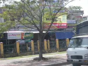 Bức ảnh ghi lại khung cảnh một con đường ở khu vực đô thị hoặc thị trấn tại Việt Nam. Ở tiền cảnh là một cây xanh lớn và một phần vỉa hè. Bên phải là phần đầu của một chiếc xe tải màu trắng. Phía sau cây là một hàng rào với các cột trụ màu vàng và song sắt màu xanh lá, bên trong có các cửa hàng và một vài chiếc xe máy đang đỗ. Các tòa nhà ở hậu cảnh là các cửa hàng kinh doanh san sát nhau, với nhiều biển hiệu quảng cáo.。Các thông tin văn bản có thể nhận dạng bao gồm:。- Biển hiệu bên trái: "ĐẠI LÝ PHƯƠNG TRƯỜNG", "Chuyên mua bán phụ tùng..." và có logo của hãng Castrol.。- Biển hiệu lớn ở giữa, màu vàng và đỏ: "DER 25", "HÙNG CƯỜNG - NHÀ PHÂN PHỐI" và số điện thoại.。- Biển hiệu bên phải, phía trên: "...IỆN MÁY - ĐỘNG CƠ ĐIỆN".。Khung cảnh cho thấy đây là một khu vực thương mại sầm uất với các cửa hàng chuyên về phụ tùng, máy móc và thiết bị điện. Bức ảnh có vẻ được chụp từ một phương tiện đang di chuyển.。（日本語訳）。この写真はベトナムの都市部または町にある道路の風景を記録したものです。前景には大きな緑の木と歩道の一部があります。右側には白いトラックの先端部分があります。木の後ろには、黄色い柱と緑色の鉄格子のあるフェンスがあり、その内側には店舗と数台のバイクが駐車しています。背景の建物は、多くの広告看板が掲げられた、隣接する商店です。識別可能なテキスト情報は次のとおりです。- 左側の看板：「ĐẠI LÝ PHƯƠNG TRƯỜNG」（フオン・チュオン代理店）、「Chuyên mua bán phụ tùng...」（部品の専門売買...）とカストロール社のロゴ。- 中央の大きな黄色と赤の看板：「DER 25」、「HÙNG CƯỜNG - NHÀ PHÂN PHỐI」（フン・クオン - 販売代理店）と電話番号。- 右側、上部の看板：「...IỆN MÁY - ĐỘNG CƠ ĐIỆN」（...電機 - 電動機）。この風景は、部品、機械、電気機器を専門とする店舗が並ぶ、にぎやかな商業地域であることを示しています。この写真は移動中の乗り物から撮影されたようです。