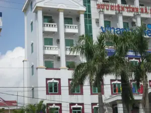 Bức ảnh chụp một tòa nhà lớn, nhiều tầng, màu trắng trong một khung cảnh đô thị vào ban ngày. Trên mặt tiền của tòa nhà có một số biển hiệu. Ở tầng trên cùng, có dòng chữ lớn màu đỏ ghi "BƯU ĐIỆN TỈNH...", phần tên tỉnh ở cuối không thể đọc được. Phía dưới là logo lớn màu xanh của "Vinaphone", một nhà cung cấp dịch vụ viễn thông. Một biển hiệu khác màu xanh cũng có thể được nhìn thấy một phần, ghi chữ "TRUNG TÂM...".。Tòa nhà có kiến trúc hiện đại với các ban công, cột trụ và nhiều cửa sổ lắp kính màu xanh lục. Một số cục nóng điều hòa được gắn bên ngoài tường. Phía trước, những cây cọ che khuất một phần của tòa nhà. Nhiều đường dây điện và cáp vắt ngang qua khung hình. Bầu trời trong xanh với những đám mây trắng. Không có hoạt động nào của con người được thể hiện rõ ràng trong ảnh.。（日本語訳）。写真は、日中の都市の風景の中にある、大きな多層階の白い建物を写しています。建物の正面にはいくつかの看板があります。最上階には、「BƯU ĐIỆN TỈNH...」と書かれた大きな赤い文字があり、末尾の省の名前の部分は読み取れません。その下には、通信サービスプロバイダーである「Vinaphone」の大きな青いロゴがあります。「TRUNG TÂM...」と書かれた別の青い看板も一部見えます。建物は近代的な建築様式で、バルコニー、柱、そして緑色のガラスがはめ込まれた多くの窓があります。いくつかのエアコンの室外機が壁の外側に取り付けられています。前面では、ヤシの木が建物の一部を覆い隠しています。多くの電線やケーブルがフレームを横切っています。空は青く澄んでおり、白い雲が浮かんでいます。写真には、人間の活動ははっきりと写っていません。