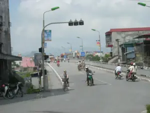 Đây là khung cảnh trên một con đường có cầu ở một khu vực đô thị tại Việt Nam. Nhiều người đang tham gia giao thông, chủ yếu bằng xe máy (xe gắn máy). Hầu hết người đi xe máy đều đội mũ bảo hiểm. Có một người đang đi xe đạp. Hai bên đường là các tòa nhà, một số trông đã cũ. Dọc theo con đường có các cột đèn đường và cột đèn tín hiệu giao thông. Có các biểu ngữ treo trên cột đèn nhưng chữ viết trên đó không thể đọc rõ. Ở phía xa, có thể nhìn thấy những dãy núi dưới bầu trời nhiều mây.。（日本語訳）。これはベトナムのある都市部にある、橋のある道路の光景です。多くの人々が、主にバイク（原動機付自転車）で交通に参加しています。バイクに乗る人のほとんどはヘルメットを着用しています。自転車に乗っている人が一人います。道路の両側には建物があり、そのうちのいくつかは古く見えます。道路に沿って街灯や信号機があります。街灯には横断幕が掲げられていますが、そこに書かれた文字ははっきりと読むことができません。遠くには、曇り空の下に山々が見えます。