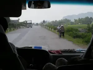 Bức ảnh được chụp từ bên trong một chiếc ô tô đang di chuyển, nhìn ra phía trước qua kính chắn gió. Khung cảnh là một con đường nông thôn ở Việt Nam, có thể là ở khu vực Ninh Bình. Con đường trải nhựa, hai bên có cọc tiêu trắng. Ở phía xa, có một chiếc xe tải và các phương tiện khác đang lưu thông. Bên phải đường, hai người dân địa phương đang đi bộ; một người phụ nữ đội nón lá truyền thống và một người đàn ông đang dắt một chiếc xe đạp. Bao quanh con đường là những cánh đồng lúa xanh và cây cối, với những dãy núi mờ ảo ở phía chân trời. Bên trong xe, có thể thấy một phần bảng điều khiển, gương chiếu hậu phản chiếu hình ảnh một người đeo kính râm, và bóng của những người ngồi trong xe. Không có văn bản hoặc chữ viết nào có thể đọc được trong ảnh.。（日本語訳）。写真は移動中の車内から、フロントガラス越しに前方を向いて撮影されたものです。風景はベトナムの田舎道で、おそらくニンビン地方のものでしょう。道路は舗装されており、両側には白い標識杭があります。遠くには、トラックやその他の車両が走行しています。道路の右側では、地元の人々が2人歩いています。伝統的なノンラーをかぶった女性と、自転車を押している男性です。道路の周りには緑の田んぼや木々が広がり、地平線にはかすんだ山脈が見えます。車内には、ダッシュボードの一部、サングラスをかけた人物の姿を映すバックミラー、そして車内に座っている人々の影が見えます。写真には読み取れるテキストや文字はありません。