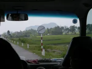 Bức ảnh được chụp từ bên trong một chiếc ô tô đang di chuyển, nhìn ra ngoài qua kính chắn gió. Khung cảnh bên ngoài là một con đường nông thôn ở Việt Nam, có khả năng là ở khu vực Ninh Bình. Bên cạnh con đường là một cánh đồng lúa xanh rộng lớn. Phía xa là những dãy núi đá vôi và cây cối. Ven đường có các cọc tiêu sơn sọc trắng đỏ và một biển báo giao thông hình tròn, nền trắng với các vạch đen chéo, báo hiệu "hết mọi lệnh cấm". Bên trong xe, có thể thấy gương chiếu hậu phản chiếu hình ảnh một người đeo kính râm, một phần ghế ngồi và một chai nước trên bảng điều khiển. Hoạt động chính là di chuyển bằng xe ô tô qua vùng nông thôn.。（日本語訳）。写真は移動中の車内から、フロントガラス越しに外を見て撮影されたものです。外の風景はベトナムの田舎道で、おそらくニンビン地方でしょう。道の脇には広大な緑の田んぼが広がっています。遠くには石灰岩の山々と木々が見えます。道端には赤と白の縞模様に塗られたデリニエーターポストと、「すべての禁止事項の終わり」を意味する、白地に黒の斜線が入った円形の交通標識があります。車内では、バックミラーにサングラスをかけた人物、座席の一部、ダッシュボードの上の水のボトルが映っているのが見えます。主な活動は、車で田園地帯を移動することです。