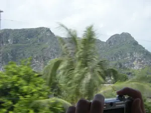 Bức ảnh được chụp từ một phương tiện đang di chuyển, có thể là ô tô hoặc tàu hỏa, thể hiện qua sự mờ nhòe của các vật thể ở tiền cảnh. Ở hậu cảnh là một dãy núi đá vôi lớn, được bao phủ bởi thảm thực vật xanh tươi. Ở tiền cảnh, mặc dù bị mờ, có thể nhận ra một cây cọ hoặc cây dừa và các bụi cây xanh khác. Ở góc dưới bên phải của bức ảnh, một người đang dùng hai tay cầm một chiếc máy ảnh kỹ thuật số màu bạc để chụp lại khung cảnh núi non. Có một vài đường dây điện chạy ngang qua bầu trời u ám, nhiều mây. Không có văn bản hay chữ viết nào có thể đọc được trong ảnh.。（日本語訳）。写真は、前景の物体がぼやけていることからわかるように、自動車か列車かもしれない移動中の乗り物から撮影されたものです。背景には、青々とした植物に覆われた大きな石灰岩の山脈があります。前景には、ぼやけてはいますが、ヤシの木かココヤシの木、そしてその他の緑の茂みが認識できます。写真の右下隅では、一人の人が銀色のデジタルカメラを両手で持ち、山の風景を撮影しています。曇り空を数本の電線が横切っています。写真の中には、読み取れるテキストや文字はありません。