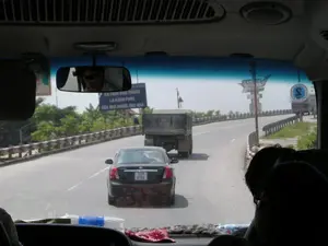 Bức ảnh được chụp từ bên trong một chiếc xe ô tô, có thể là xe khách, nhìn ra quang cảnh giao thông phía trước qua kính chắn gió. Khung cảnh cho thấy một con đường nhựa, có vẻ là một cây cầu hoặc đường trên cao, với lan can bảo vệ ở hai bên. Trên đường, có nhiều phương tiện đang di chuyển, bao gồm một chiếc ô tô con màu đen và một chiếc xe tải lớn ở phía trước. Bên phải đường có một tấm biển lớn màu xanh dương với khẩu hiệu tuyên truyền về an toàn giao thông: "AN TOÀN GIAO THÔNG LÀ HẠNH PHÚC CỦA MỌI NGƯỜI, MỌI NHÀ". Ở phía xa bên phải, có thể thấy một công trình kiến trúc mang tính biểu tượng và một chiếc xe khách khác. Bên trong xe, gương chiếu hậu phản chiếu hình ảnh một người đeo kính râm. Trên tấm chắn nắng có dán một bức ảnh chân dung. Trên bảng điều khiển có một chai nước và có thể thấy bóng của những hành khách khác trong xe.。（日本語訳）。写真は自動車、おそらくはバスの車内から、フロントガラス越しに前方の交通風景を撮影したものです。道路上には、前方に黒い乗用車1台と大型トラック1台を含む、多くの車両が走行しています。道路の右側には、「交通安全は、すべての人、すべての家庭の幸福です」という交通安全に関するスローガンが書かれた大きな青い看板があります。右側の遠方には、象徴的な建築物と別のバスが見えます。車内では、バックミラーにサングラスをかけた人の姿が映っています。サンバイザーには肖像写真が貼られています。