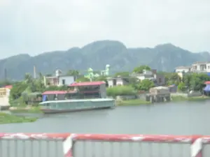 Bức ảnh chụp một khung cảnh ven sông với những dãy núi đá vôi đặc trưng ở phía xa, có khả năng là tại khu vực Ninh Bình. Ở tiền cảnh là lan can cầu màu đỏ và trắng bị mờ. Dưới sông, một chiếc tàu du lịch lớn hai tầng, sơn màu xanh và có mái đỏ, đang neo đậu sát bờ. Bờ sông có nhiều cây cối và các công trình nhà ở. Trong số các tòa nhà, có một công trình kiến trúc màu xanh lá cây với mái vòm, có thể là một nhà thờ. Phía sau là dãy núi đá vôi hùng vĩ. Toàn bộ khung cảnh khá mờ, có thể do được chụp khi đang di chuyển. Không có văn bản hay biển hiệu nào có thể nhận dạng được.。（日本語訳）。写真は、遠くに特徴的な石灰岩の山脈がある川沿いの風景を写しており、おそらくニンビン地方のものである。前景には、ぼやけた赤と白の橋の欄干がある。川には、青く塗られ赤い屋根を持つ2階建ての大きな観光船が1隻、岸辺に停泊している。川岸には多くの木々や住宅の建物がある。建物の中には、ドーム型の屋根を持つ緑色の建築物があり、おそらく教会であろう。後ろには雄大な石灰岩の山脈がある。風景全体はかなりぼやけており、移動中に撮影されたためかもしれない。認識できる文字や看板はない。