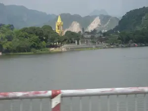 Bức ảnh ghi lại một khung cảnh ven sông ở một vùng nông thôn, có thể là ở Ninh Bình, được chụp từ trên một cây cầu. Ở tiền cảnh là một phần lan can cầu màu trắng và đỏ. Phía bên kia bờ sông là một khu dân cư với những ngôi nhà nằm xen lẫn giữa những lùm cây xanh tươi. Nổi bật trong khung cảnh là một tháp chuông nhà thờ Công giáo có kiến trúc kiểu Gothic, được sơn màu vàng. Phía sau làng mạc và nhà thờ là những dãy núi đá vôi hùng vĩ, một vài ngọn núi có dấu hiệu bị khai thác đá, để lộ ra những vách đá màu trắng. Dọc theo bờ sông, có một vài chiếc thuyền nhỏ đang neo đậu. Khung cảnh tổng thể yên bình, không có hoạt động rõ rệt của con người.。（日本語訳）。写真は、おそらくニンビン省の、とある田舎の川沿いの風景を捉えたもので、橋の上から撮影されたものです。川の対岸には住宅地があり、家々が青々とした木々の間に点在しています。風景の中でひときわ目立っているのは、ゴシック様式の建築で、黄色に塗られたカトリック教会の鐘楼です。村と教会の後ろには雄大な石灰岩の山々が連なっており、いくつかの山には石を採掘した形跡があり、白い崖が露出しています。川岸に沿って、数隻の小さな船が停泊しています。全体的な風景は穏やかで、目立った人の活動は見られません。