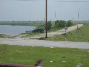 Bức ảnh ghi lại một khung cảnh nông thôn, có khả năng được chụp từ một phương tiện đang di chuyển. Cảnh vật bao gồm một con đường đất chạy dọc theo một hồ nước hoặc một khúc sông rộng. Con đường này uốn lượn và có một biển báo giao thông hình tròn cắm bên lề, tuy nhiên nội dung trên biển báo không thể nhận dạng được do hình ảnh bị mờ. Trên mặt nước ở phía xa, có thể thấy một chiếc thuyền hoặc sà lan. Cảnh quan xung quanh là những bãi cỏ xanh, một vài bụi cây và các cột điện. Phía xa hơn là những cánh đồng rộng lớn. Ở tiền cảnh, có một phần của con đường nhựa và bãi cỏ ven đường có một ít rác.。（日本語訳）。写真は田舎の風景を記録しており、移動中の乗り物から撮影された可能性があります。風景には、湖または川幅の広い川の一部分に沿って走る一本の未舗装の道が含まれています。この道は曲がりくねっており、道端には円形の交通標識が立てられていますが、画像がぼやけているため標識の内容は識別できません。遠くの水面には、一隻のボートまたははしけが見えます。周囲の景観は、緑の草地、いくつかの茂み、そして電柱です。さらに遠くには広大な田畑が広がっています。前景には、舗装道路の一部と、少しゴミのある道端の草地があります。