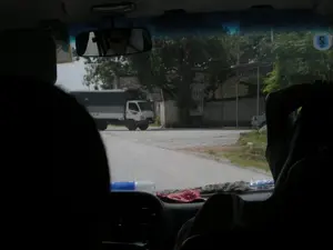 Bức ảnh được chụp từ bên trong một chiếc ô tô, nhìn ra ngoài qua kính chắn gió. Khung cảnh bên trong xe bị tối, chỉ thấy bóng của người ngồi và một phần bảng điều khiển. Gương chiếu hậu phản chiếu khuôn mặt của người lái xe đang đeo kính râm. Bên ngoài, trên một con đường nhựa, có một chiếc xe tải màu trắng với thùng xe phủ bạt đang đỗ. Phía xa là cây cối và một ngôi nhà. Có một tấm biển hiệu dựng bên đường, trên đó có các chữ cái nhưng chỉ có thể nhận ra một phần là "CƠM PHỞ", gợi ý rằng có một quán ăn ở gần đó. Toàn cảnh cho thấy một con đường ở vùng nông thôn hoặc ngoại ô Việt Nam.。（日本語訳）。写真は車内から、フロントガラス越しに外を見て撮影されたものです。車内は暗く、座っている人の影とダッシュボードの一部だけが見えます。バックミラーにはサングラスをかけた運転手の顔が映っています。外の舗装道路には、荷台に幌をかけた白いトラックが1台停まっています。遠くには木々と一軒の家があります。道端に看板が立てられており、そこには文字が書かれていますが、「CƠM PHỞ（コム・フォー）」という部分しか認識できず、近くに食堂があることを示唆しています。全景は、ベトナムの田舎または郊外の道を示しています。