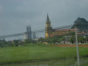 Bức ảnh ghi lại một khung cảnh vùng nông thôn, có khả năng là ở khu vực Ninh Bình, với sự pha trộn giữa các yếu tố nông nghiệp, tôn giáo và công nghiệp. Ở tiền cảnh là một cánh đồng lúa xanh mướt và một phần ao nước. Nổi bật ở giữa là một nhà thờ Công giáo lớn màu vàng với tháp chuông cao theo kiến trúc Gô-tích và mái ngói đỏ. Phía sau nhà thờ, bên phải là một ngọn núi đá vôi, trên đỉnh có một cây thánh giá. Ở phía xa bên trái là một khu công nghiệp lớn, dường như là một nhà máy xi măng với các xi-lô và cấu trúc cao tầng. Bầu trời u ám, có nhiều mây. Nhiều đường dây điện chạy ngang qua khung cảnh.。（日本語訳）。写真は、おそらくニンビン地方の田舎の風景を捉えたもので、農業、宗教、工業の要素が混在している。前景には、青々とした水田と池の一部がある。中央に目立つのは、ゴシック建築の高い鐘楼と赤い瓦屋根を持つ、黄色い大きなカトリック教会である。教会の後ろ、右側には石灰岩の山があり、その頂上には十字架が立っている。遠くの左側には大きな工業地帯があり、サイロや高層構造物を持つセメント工場のようだ。空はどんよりとしており、雲が多い。多くの電線が風景を横切っている。