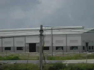 Hình ảnh chụp một tòa nhà lớn, có khả năng là một nhà máy hoặc nhà kho công nghiệp. Tòa nhà có cấu trúc hiện đại với tường và mái được làm bằng tôn kim loại màu trắng, phần móng dưới được xây bằng bê tông. Một ống thông gió lớn hình trụ chạy dọc theo nóc nhà. Tòa nhà dường như đang trong giai đoạn xây dựng hoặc vừa mới hoàn thành, với khu đất xung quanh còn ngổn ngang và được rào lại sơ sài. Qua các ô cửa sổ ở tầng trệt, có thể thấy một vài chiếc xe ô tô màu trắng đang đỗ bên trong. Ở tiền cảnh, có một cột điện với nhiều dây điện và dây cáp vắt ngang, che khuất một phần của tòa nhà. Bầu trời u ám và có nhiều mây. Không có bất kỳ văn bản hay biển hiệu nào có thể đọc được.。（日本語訳）。大きな建物の写真で、工場または工業用倉庫である可能性があります。建物は現代的な構造で、壁と屋根は白色の金属製トタンでできており、下部の基礎はコンクリートで造られています。大きな円筒形の換気ダクトが屋根に沿って走っています。建物は建設中か、あるいは完成したばかりのようで、周囲の土地はまだ散らかっており、粗末な柵で囲われています。1階の窓からは、中に数台の白い自動車が駐車しているのが見えます。前景には、多くの電線やケーブルが横切っている電柱があり、建物の一部を覆い隠しています。空はどんよりと曇っています。読み取れる文字や看板は一切ありません。