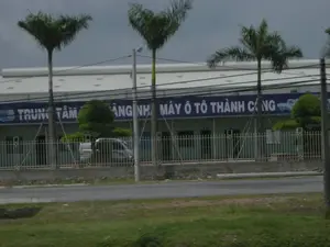 Bức ảnh chụp một tòa nhà lớn, có vẻ là một nhà máy hoặc trung tâm dịch vụ ô tô. Trên mặt tiền của tòa nhà có một tấm biển hiệu lớn màu xanh lam với dòng chữ trắng. Dòng chữ có thể đọc được là "TRUNG TÂM ... MÁY Ô TÔ THÀNH CÔNG", một phần của văn bản bị che khuất bởi cây cối và cột điện. Khuôn viên được bao quanh bởi một hàng rào kim loại. Phía trước tòa nhà có trồng một số cây cọ và cây cảnh được cắt tỉa. Một chiếc xe van màu trắng đang đỗ bên trong hàng rào. Phía trước là một con đường nhựa và một dải cỏ. Bầu trời u ám. Không có hoạt động nào của con người được quan sát thấy.。（日本語訳）。写真には大きな建物が写っており、工場か自動車サービスセンターのようです。建物の正面には、白い文字が書かれた大きな青い看板があります。読み取れる文字は「TRUNG TÂM ... MÁY Ô TÔ THÀNH CÔNG」で、テキストの一部は木々や電柱で隠されています。敷地は金属製のフェンスで囲まれています。建物の前には、数本のヤシの木と刈り込まれた観賞用の木が植えられています。フェンスの内側には白いバンが1台駐車しています。手前にはアスファルトの道路と芝生の帯があります。空は曇っています。人の活動は見られません。