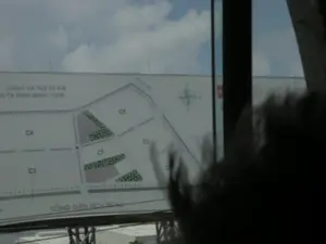 Bức ảnh chụp một tấm biển quảng cáo lớn bên đường, nhìn từ bên trong một phương tiện đang di chuyển. Tấm biển này giới thiệu sơ đồ quy hoạch mặt bằng của một dự án. Các thông tin bằng chữ có thể đọc được trên biển quảng cáo bao gồm: 'CÁCH HÀ NỘI 82 KM', 'ĐẾN TX NINH BÌNH 11KM' (chữ 'ĐẾN TX' không rõ nét nhưng có thể suy đoán), và 'TỔNG DIỆN TÍCH 25 ha'. Sơ đồ trên biển thể hiện các lô đất được phân chia và ký hiệu là C1, C2, C3, C4, C5, cùng với các khu vực được tô màu xanh lá cây (có thể là khu cây xanh) và các khu vực màu khác. Trên sơ đồ còn có một hoa tiêu chỉ phương hướng. Phía trên tấm biển là bầu trời có mây. Ở tiền cảnh bên phải là bóng mờ của đầu một người.。（日本語訳）。移動中の乗り物の中から見た、道端の大きな広告看板を撮影した写真。この看板は、あるプロジェクトの平面計画図を紹介しています。看板で読み取れる文字情報には、「ハノイまで82KM」、「ニンビン市まで11KM」（「ĐẾN TX」の文字は不鮮明ですが推測可能です）、そして「総面積25ヘクタール」が含まれます。看板上の図は、C1、C2、C3、C4、C5と記号が付けられた分割された区画、緑色で塗られたエリア（おそらく緑地）、およびその他の色のエリアを示しています。図には方位を示すコンパスローズもあります。看板の上は雲のある空です。前景の右側には、人の頭のぼやけた影があります。