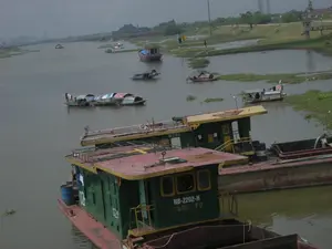 Bức ảnh chụp một khung cảnh trên một con sông hoặc kênh đào ở khu vực Ninh Bình. Bầu trời u ám và mặt nước có màu xám đục.。Trong ảnh có rất nhiều tàu thuyền. Ở phía trước là hai chiếc sà lan chở hàng lớn, có cabin điều khiển. Cabin được sơn màu xanh lá cây và vàng, còn thân tàu màu đỏ. Trên cabin của chiếc sà lan gần nhất có thể đọc được các ký tự: "NB-2202-H" và bên dưới là "116T / Y.Đ". Chiếc sà lan ngay phía sau nó có số hiệu "NB-2718-H". "NB" là biển đăng ký của tỉnh Ninh Bình, cho thấy đây là những phương tiện hoạt động tại địa phương.。Ở phía xa hơn, có nhiều thuyền nhỏ hơn đang neo đậu hoặc di chuyển trên sông. Đáng chú ý là một dãy gồm khoảng bốn hoặc năm chiếc thuyền nhỏ có mái che tạm bợ bằng bạt và các vật liệu khác, được buộc lại với nhau, trông giống như một khu nhà ở nổi của một gia đình. Có thể thấy quần áo đang được phơi trên một trong những chiếc thuyền này. Ngoài ra còn có các thuyền gỗ và thuyền máy nhỏ khác nằm rải rác.。Hai bên bờ sông là đất và cỏ xanh. Ở phía xa có thể thấy các cột điện cao thế và một vài công trình xây dựng. Trên mặt nước có những đám bèo tây (lục bình) trôi nổi. Khung cảnh cho thấy một hoạt động vận tải đường thủy và cuộc sống sinh hoạt thường ngày trên sông.。（日本語訳）。この写真は、ニンビン地域の川または運河の風景を撮影したものです。空は曇っており、水面は濁った灰色をしています。手前には、操縦室のある2隻の大きな貨物船（はしけ）があります。操縦室は緑と黄色に塗られており、船体は赤色です。最も手前のはしけの操縦室には、「NB-2202-H」という文字が読み取れ、その下には「116T / Y.Đ」と書かれています。そのすぐ後ろのはしけには、「NB-2718-H」という番号が付いています。「NB」はニンビン省の登録標識であり、これらが地元で活動している船であることを示しています。さらに遠くには、より多くの小さな船が川に停泊したり移動したりしています。これらの船の一隻には、洗濯物が干されているのが見えます。その他にも、木造船や小型のモーターボートが点在しています。川の両岸は土と緑の草です。遠くには高圧電線の鉄塔やいくつかの建造物が見えます。水面にはホテイアオイ（lục bình）の群れが浮かんでいます。この光景は、水上輸送の活動と川での日常生活を示しています。