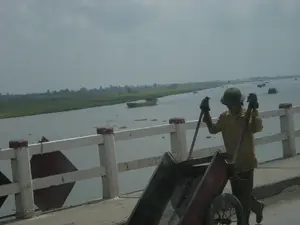 Bức ảnh chụp cảnh một người lao động đang đẩy một chiếc xe đẩy tay trên một cây cầu bê tông. Người này mặc đồng phục lao động màu vàng, đội mũ cối màu xanh và đeo găng tay. Cây cầu có lan can màu trắng với các cột trụ có đỉnh sơn đỏ, bắc qua một con sông rộng. Dưới sông, có một vài chiếc tàu và sà lan đang di chuyển. Phía xa là bờ sông với những cánh đồng xanh. Có một biển báo hình thoi màu đỏ gắn trên lan can nhưng không thể đọc rõ nội dung.。（日本語訳）。コンクリートの橋の上で、一人の労働者が手押し車を押している様子の写真です。この人物は黄色の作業服を着て、緑色のヘルメットをかぶり、手袋をしています。橋には白い手すりがあり、その支柱の先端は赤く塗られていて、広い川に架かっています。川には、数隻の船とはしけが移動しています。遠くには、緑の野原が広がる川岸があります。手すりには赤い菱形の標識が取り付けられていますが、内容ははっきりと読み取れません。