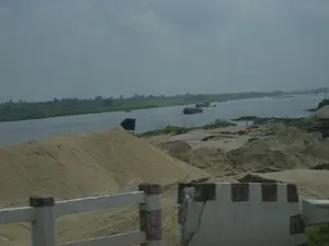 Bức ảnh chụp một khung cảnh ven sông rộng lớn, có thể là ở khu vực Ninh Bình. Ở tiền cảnh, có những đống cát khổng lồ được chất cao bên bờ, cho thấy hoạt động khai thác hoặc tập kết vật liệu xây dựng. Một hàng rào chắn bê tông, có thể là của một con đường hoặc cây cầu, xuất hiện ở gần nhất. Trên mặt sông, nhiều tàu chở hàng (sà lan) đang hoạt động, có khả năng đang vận chuyển cát. Bờ sông phía xa là một dải đất xanh với cây cối. Có một biển báo màu đen hình mũi tên gần bờ sông nhưng không có chữ. Bầu trời có nhiều mây và khá u ám. Toàn cảnh gợi lên một hoạt động kinh tế sôi động liên quan đến vận tải đường thủy và khai thác tài nguyên thiên nhiên.。（日本語訳）。写真は、おそらくニンビン地域の、広大な川沿いの風景を写しています。前景には、岸辺に高く積まれた巨大な砂の山があり、採掘または建設資材の集積活動を示唆しています。おそらく道路か橋のものであるコンクリートの防護柵が、最も手前に見えます。川面では、多くの貨物船（はしけ）が活動しており、おそらく砂を運んでいます。遠くの川岸は、木々のある緑の地帯です。川岸の近くに黒い矢印の形をした標識がありますが、文字はありません。空は雲が多く、かなり曇っています。全景は、水上輸送と天然資源の採掘に関連する活発な経済活動を想起させます。