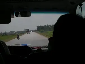Bức ảnh được chụp từ bên trong một chiếc ô tô, nhìn ra ngoài qua kính chắn gió về một con đường ở vùng nông thôn. Trên con đường nhựa, có một người đang đi xe máy đội mũ bảo hiểm màu đỏ và ở phía xa hơn là một chiếc xe buýt hoặc xe tải. Hai bên đường là cánh đồng xanh và các cọc tiêu bê tông. Ở phía xa có một tấm biển quảng cáo lớn nhưng không thể đọc được nội dung. Tiền cảnh là bóng của một người ngồi trong xe và một phần bảng điều khiển của ô tô. Gương chiếu hậu phản chiếu hình ảnh một người đang đeo kính râm.。（日本語訳）。写真は車内から撮影されたもので、フロントガラス越しに田舎の道が見えます。アスファルトの道路には、赤いヘルメットをかぶった人がバイクに乗っており、さらに遠くにはバスかトラックがいます。道路の両側には緑の野原とコンクリートの杭があります。遠くには大きな広告看板がありますが、内容は読み取れません。前景は、車内に座っている人の影と、自動車のダッシュボードの一部です。バックミラーには、サングラスをかけている人の姿が映っています。