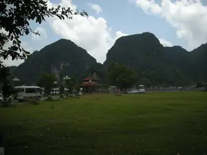 Bức ảnh chụp một khung cảnh ngoài trời tại một khu vực có nhiều núi đá vôi, có khả năng là một điểm du lịch hoặc bãi đỗ xe ở Ninh Bình. Tiền cảnh là một bãi cỏ xanh rộng. Có một con đường nhỏ nơi một chiếc xe buýt nhỏ màu trắng đang chạy. Gần đó có một ngôi nhà lầu nhỏ kiểu kiến trúc truyền thống với mái ngói đỏ, có thể là nhà chờ hoặc vọng lâu. Dọc đường có các cột đèn đường. Ở phía xa hơn là một bãi đỗ xe với nhiều ô tô và xe buýt du lịch khác đang đỗ. Phông nền nổi bật là những dãy núi đá vôi hùng vĩ, được bao phủ bởi cây cối xanh tốt. Bầu trời trong xanh và có mây. Một vài người xuất hiện trong ảnh, có vẻ là khách du lịch đang dừng chân tham quan. Trên thân chiếc xe buýt nhỏ màu trắng có chữ viết nhưng không thể đọc rõ.。（日本語訳）。写真は、多くの石灰岩の山がある屋外の風景を撮影したもので、おそらくニンビンの観光地または駐車場である。前景には広い緑の芝生が広がっている。小さな道があり、そこを一台の白い小型バスが走っている。その近くには、伝統的な建築様式の赤い瓦屋根を持つ小さな建物があり、待合所か展望台かもしれない。道沿いには街灯が立っている。さらに奥には駐車場があり、多くの乗用車や他の観光バスが駐車している。背景には、青々とした木々に覆われた雄大な石灰岩の山々が際立っている。空は青く澄んでおり、雲がある。写真には数人の人物が写っており、立ち寄って観光している旅行者のようだ。白い小型バスの車体には文字が書かれているが、はっきりと読み取ることはできない。