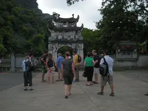 Trên cổng tam quan bằng đá có khắc bốn chữ Hán: 鎮靖門北 (Trấn Tĩnh Môn Bắc).。Bối cảnh là một khoảng sân bê tông ở lối vào của một khu di tích lịch sử hoặc đền chùa. Một nhóm du khách khoảng 10 người đang đứng tập trung trước cổng. Họ mặc trang phục thường ngày, thoải mái cho việc đi du lịch (áo phông, quần soóc, dép) và một số người đeo ba lô. Có vẻ như họ đang chuẩn bị vào tham quan hoặc đang lắng nghe hướng dẫn.。Công trình nổi bật là chiếc cổng tam quan cổ kính, hai tầng, bằng đá với mái ngói cong theo kiến trúc truyền thống. Phía sau và bên trái cổng là một ngọn núi đá vôi được bao phủ bởi thảm thực vật xanh tươi, rậm rạp. Bên phải cổng là một bức tường và một công trình nhỏ có mái che, bên trong dường như có một tấm bia đá. Cây cối xanh tốt mọc xung quanh khu vực.。（日本語訳）。石造りの三関門には「鎮靖門北（Trấn Tĩnh Môn Bắc）」という四つの漢字が刻まれている。背景は、史跡または寺院の入り口にあるコンクリートの庭である。約10人の観光客のグループが門の前に集まって立っている。彼らは旅行に適した普段着（Tシャツ、ショートパンツ、サンダル）を着用しており、何人かはバックパックを背負っている。彼らは見学に入る準備をしているか、ガイドの説明を聞いているようだ。目立つ建造物は、伝統建築様式に沿った反りのある瓦屋根を持つ、石造りの古風な二層の三関門である。門の後方と左側には、青々とした鬱蒼とした植物に覆われた石灰岩の山がある。門の右側には壁と屋根付きの小さな建造物があり、その中には石碑があるようだ。