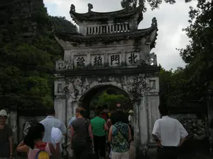 Bức ảnh chụp một cổng thành cổ bằng đá, mang đậm nét kiến trúc truyền thống với mái ngói cong hai tầng. Cổng đã cũ, nhuốm màu thời gian. Phía trên vòm cổng có khắc bốn chữ Hán lớn, đọc từ phải sang trái là 北門鋪鎮 (Bắc Môn Phố Trấn). Bối cảnh phía sau là một ngọn núi đá vôi cao, cây cối um tùm bao phủ, một cảnh quan đặc trưng của vùng Ninh Bình. Hoạt động chính trong ảnh là một nhóm người, có vẻ là du khách, đang tụ tập trước cổng. Họ mặc trang phục thường ngày, thoải mái, cho thấy họ đang đi tham quan một địa điểm du lịch lịch sử. Một số người đang tiến vào qua cổng, trong khi những người khác đứng quan sát.。（日本語訳）。写真は石造りの古い城門を写しており、二層の反りのある瓦屋根を持つ伝統的な建築様式が色濃く表れている。門は古く、時の色が染みついている。門のアーチの上には大きな漢字四文字が刻まれており、右から左に「北門鋪鎮」（バック・モン・フォー・チャン）と読む。背景には、木々が生い茂る高い石灰岩の山があり、ニンビン地方特有の景観である。写真の中の主な活動は、観光客と思われる一団が門の前に集まっていることである。彼らは普段着の楽な服装をしており、歴史的な観光地を訪れていることを示している。何人かは門を通り抜けて中へ進んでおり、他の人々は立って観察している。