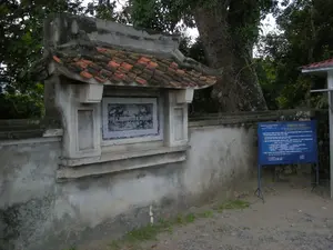 Đây là hình ảnh chụp tại một khu di tích lịch sử.。**1. Chữ viết:**。- Trên một tấm bia đá màu đen được gắn vào tường có khắc dòng chữ: "ĐỀN THỜ VUA ĐINH TIÊN HOÀNG".。- Bên phải là một bảng thông báo màu xanh dương có tiêu đề "THÔNG BÁO". Nội dung trên bảng, do "BAN QUẢN LÝ DI TÍCH CỐ ĐÔ HOA LƯ" ban hành, thông báo về giá vé tham quan đền thờ Vua Đinh - Vua Lê cho du khách, bao gồm giá vé cho người lớn, trẻ em và giá vé gửi xe máy, ô tô. Có thể đọc được các mức giá như 10.000đ và 2.000đ. Bảng thông báo có ghi ngày tháng, dường như là "ngày 01 tháng 08 năm 2008".。**2. Bối cảnh và Hoạt động:**。- **Loại địa điểm:** Đây là khu vực bên ngoài của Đền thờ Vua Đinh Tiên Hoàng, thuộc Quần thể di tích Cố đô Hoa Lư tại Ninh Bình.。- **Vật thể trong ảnh:** Bức ảnh cho thấy một bức tường bê tông đã cũ. Trên tường có một kiến trúc nhỏ mang phong cách truyền thống với mái cong lợp ngói âm dương màu đỏ cam. Bên dưới mái là tấm bia đá kể trên. Phía sau bức tường là cây cối xanh tươi và một thân cây cổ thụ lớn. Mặt đất là nền đất, có một ít cỏ mọc.。- **Hoạt động:** Không có người xuất hiện trong ảnh, do đó không có hoạt động nào đang diễn ra. Đây là một khung cảnh tĩnh tại lối vào khu di tích.。（日本語訳）。これは歴史遺跡で撮影された写真です。**1. 文字:**。- 壁に取り付けられた黒い石碑に「ĐỀN THỜ VUA ĐINH TIÊN HOÀNG」（丁先皇祠）という文字が刻まれています。- 右側には「THÔNG BÁO」（お知らせ）というタイトルの青い掲示板があります。掲示板の内容は、「BAN QUẢN LÝ DI TÍCH CỐ ĐÔ HOA LƯ」（ホアルー古都遺跡管理委員会）が発行したもので、丁王祠・黎王祠を訪れる観光客向けの参観料金について通知しており、大人、子供の料金、およびバイク、自動車の駐車料金が含まれています。10,000ドンや2,000ドンといった料金が読み取れます。掲示板には日付が記載されており、「2008年08月01日」のようです。**2. 背景と活動:**。- **場所の種類:** これは、ニンビン省にあるホアルー古都遺跡群に属する、丁先皇祠の外側のエリアです。壁の上には、赤橙色の陰陽瓦で葺かれた反りのある屋根を持つ、伝統的な様式の小さな建造物があります。屋根の下には前述の石碑があります。壁の後ろには青々とした木々と大きな古木の幹があります。地面は土で、少し草が生えています。- **活動:** 写真には人が写っていないため、何の活動も行われていません。これは遺跡の入り口での静かな光景です。