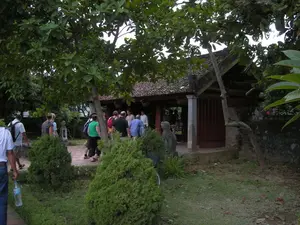 Bối cảnh là một khu di tích lịch sử hoặc một ngôi đền cổ, có thể ở Ninh Bình. Một nhóm du khách đang đi bộ trên một lối đi bằng gạch trong một khu vườn để vào tham quan một công trình kiến trúc truyền thống. Công trình này có mái ngói, cột trụ và cửa gỗ. Xung quanh là không gian sân vườn với nhiều cây xanh lớn, cây bụi trang trí và các bức tượng đá. Nhóm du khách, bao gồm cả nam và nữ, mặc trang phục đời thường và một số người mang ba lô. Hoạt động chính là tham quan, khám phá di tích. Không có văn bản hay chữ viết nào có thể đọc được trong hình ảnh.。（日本語訳）。背景は歴史的遺跡または古い寺院で、おそらくニンビン省にある。観光客の一団が、伝統的な建築物を見学するため、庭園内のレンガ敷きの小道を歩いている。この建物には瓦屋根、柱、そして木製の扉がある。周囲は庭園空間で、多くの大きな緑の木々、装飾用の低木、そして石像がある。男女を含む観光客の一団は、普段着を着用しており、何人かはバックパックを背負っている。主な活動は遺跡の見学、探索である。画像の中には判読できるテキストや文字はない。