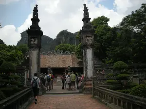 Bối cảnh là một khu di tích lịch sử hoặc đền chùa ở Ninh Bình. Lối vào là một cây cầu lát gạch đỏ dẫn đến một cổng được tạo bởi hai cột đá lớn, cổ kính. Trên thân cột đá có khắc chữ Hán hoặc chữ Nôm nhưng không thể đọc rõ. Trên đỉnh mỗi cột đá có một bức tượng linh vật, có thể là con Nghê. Một nhóm du khách đang đi vào tham quan, một số người đang đứng tụ tập ở khu vực cổng. Phía sau cổng là một công trình kiến trúc cổ với mái ngói màu nâu. Khung cảnh xung quanh được bao bọc bởi cây cối xanh tốt, cây cảnh được cắt tỉa và những dãy núi đá vôi hùng vĩ ở phía xa.。（日本語訳）。背景はニンビンにある歴史的遺跡または寺院です。入り口は、赤レンガで舗装された橋で、二本の大きな古風な石柱で作られた門に通じています。石柱の本体には漢字またはチュノムが刻まれていますが、はっきりと読むことはできません。各石柱の上には、おそらくゲー（Nghê）という霊獣の像があります。観光客のグループが見学のために入ってきており、何人かは門のあたりに集まって立っています。門の後ろには、茶色の瓦屋根を持つ古い建築物があります。周囲の風景は、青々とした木々、手入れされた盆栽、そして遠くにある雄大な石灰岩の山々に囲まれています。