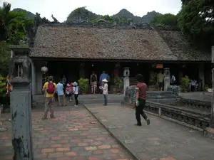 Bối cảnh là một khu di tích lịch sử hoặc một ngôi đền cổ, có thể ở Ninh Bình, với những ngọn núi đá vôi đặc trưng ở phía sau. Tòa nhà chính có kiến trúc truyền thống Việt Nam với mái ngói âm dương màu nâu đỏ và trên nóc mái được trang trí công phu bằng các hình tượng điêu khắc giống như rồng. Một nhóm du khách, bao gồm cả người nước ngoài, đang tham quan địa điểm này. Một số người đang đứng và ngồi trên bậc thềm dẫn vào đền, trong khi những người khác đang đi dạo trên sân gạch phía trước. Một người đàn ông mặc áo vàng đeo ba lô màu đỏ, một người phụ nữ đội nón lá. Một người đàn ông khác mặc áo nâu đang đi bộ trên lối đi bằng gạch. Xung quanh đền có cây cối xanh tươi và các chậu cây cảnh. Có các cột đá và lan can đá trong sân. Có một chiếc đèn lồng treo dưới mái hiên. Có vẻ như có một tấm biển phía trên lối vào nhưng không thể đọc được nội dung.。（日本語訳）。背景は歴史的遺跡または古い寺院で、おそらくニンビンにあり、背後には特徴的な石灰岩の山々があります。本殿はベトナムの伝統的な建築様式で、赤褐色の陰陽瓦の屋根を持ち、屋根の上は龍のような彫刻像で精巧に装飾されています。外国人を含む観光客のグループがこの場所を訪れています。何人かは寺院に通じる階段に立ったり座ったりしており、他の人々は前のレンガ敷きの中庭を散策しています。黄色いシャツを着て赤いバックパックを背負った男性、ノンラーをかぶった女性がいます。別の茶色いシャツを着た男性がレンガの小道を歩いています。寺院の周りには緑豊かな木々や鉢植えの観葉植物があります。中庭には石の柱と石の手すりがあります。軒下に提灯が一つ吊るされています。入り口の上には看板があるようですが、内容は読み取れません。