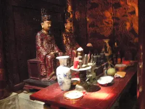 Đây là một không gian thờ cúng trong nhà, có thể là bên trong một ngôi đền hoặc chùa. Khung cảnh có ánh sáng yếu, mang lại cảm giác trang nghiêm và cổ kính. Ở phía sau có hai bức tượng ngồi, dường như là tượng vua hoặc các vị quan lại, mặc trang phục triều đình màu đỏ và vàng, đội mũ miện. Phía trước các bức tượng là một bàn thờ bằng gỗ sơn đỏ. Trên bàn thờ có bày các vật phẩm thờ cúng, bao gồm một lọ hoa lớn bằng sứ men lam, nến đỏ trong cốc, đĩa đựng lễ vật, một quả trái cây màu xanh (trông giống quả bưởi), và các đồ vật khác như lư hương. Không có chữ viết nào có thể nhận dạng được trong ảnh.。（日本語訳）。これは屋内の礼拝空間で、寺院か仏塔の内部である可能性があります。光景は薄暗く、荘厳で古風な感覚をもたらします。後方には2体の座像があり、王または高官の像のようです。赤と黄色の朝廷の衣装を身に着け、冠をかぶっています。像の前には、赤く塗られた木製の祭壇があります。祭壇の上には、青磁の大きな花瓶、コップに入った赤いろうそく、供物を入れる皿、緑色の果物（ザボンに似ています）、そして香炉などの祭祀用品が供えられています。写真の中には、認識できる文字はありません。