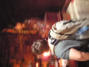 Bức ảnh rất mờ và tối, được chụp trong một không gian thiếu sáng, có thể là trong nhà vào ban đêm. Toàn bộ hình ảnh bị nhòe do chuyển động. Ở phía trước, một người đang đeo một chiếc ba lô lớn và cúi người về phía trước. Bối cảnh phía sau dường như là một quán bar hoặc nhà hàng, với các kệ chứa nhiều đồ vật trông giống như cốc thủy tinh và chai lọ đang phản chiếu ánh sáng mờ ảo, ấm áp. Có một nguồn sáng nhỏ, có thể là một ngọn nến, ở gần đầu của người đó. Do hình ảnh bị mờ và chất lượng thấp nên không thể xác định được hoạt động cụ thể hay đọc bất kỳ văn bản nào.。（日本語訳）。写真は非常にぼやけて暗く、夜間の室内など、光が少ない空間で撮影されたものです。画像全体が動きによってぶれています。前景には、大きなバックパックを背負い、前かがみになっている人物がいます。背景はバーかレストランのようで、棚にはガラスのコップや瓶のような多くの物が置かれ、ぼんやりとした暖かい光を反射しています。その人物の頭の近くに、おそらくろうそくと思われる小さな光源があります。画像がぼやけていて質が低いため、具体的な活動を特定したり、文字を読み取ったりすることはできません。