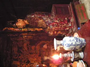 Bức ảnh chụp một không gian nội thất tối, có khả năng là bên trong một ngôi đền hoặc lăng mộ. Đối tượng chính là một bức tượng có kích thước như người thật của một người đang nằm, có thể là một vị vua hoặc một nhân vật lịch sử quan trọng. Bức tượng được đặt trên một chiếc long sàng (giường của vua) hoặc bệ thờ lớn, được sơn son thếp vàng và trang trí rất công phu. Phần thân của bệ thờ được chạm khắc các hoa văn tinh xảo, trong đó có họa tiết rồng màu vàng trên nền tối. Ở phía trước, một ngọn nến đang cháy soi sáng một phần của cảnh vật, bên cạnh đó là một chiếc bình gốm sứ hoa lam trắng. Ở phía sau, có một tấm biển màu xanh lá cây với chữ viết màu đỏ, dù hơi mờ nhưng có thể đọc được là 'ĐINH TIÊN HOÀNG'. Điều này cho thấy địa điểm có thể là đền thờ Vua Đinh Tiên Hoàng ở Hoa Lư, Ninh Bình. Khung cảnh trang trọng và các vật phẩm cho thấy đây là một nơi thờ cúng và tưởng niệm.。（日本語訳）。写真は暗い室内空間を写しており、おそらく寺院か霊廟の内部だと思われます。主な被写体は、横たわっている人物の等身大の像で、王または重要な歴史上の人物である可能性があります。その像は、金箔を施した漆塗りで非常に精巧に装飾された、大きな龍床（王の寝台）または祭壇の上に置かれています。祭壇の胴体部分には精巧な文様が彫られており、その中には暗い背景に金色の龍の模様があります。手前では、燃えているろうそくが景色の一部を照らしており、その隣には青と白の染付の陶磁器の壺があります。後方には、緑色の地に赤色の文字が書かれた看板があり、少しぼやけていますが「ĐINH TIÊN HOÀNG（丁先皇）」と読み取ることができます。このことから、この場所はニンビン省ホアルーにある丁先皇（ディン・ティエン・ホアン）の祠である可能性が示唆されます。荘厳な光景と品々は、ここが礼拝と追悼の場所であることを示しています。