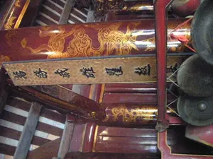 Bên trong một công trình kiến trúc cổ, có thể là một ngôi đền hoặc đình làng. Nổi bật là các xà nhà bằng gỗ được sơn son màu đỏ sẫm và trang trí bằng các họa tiết uốn lượn màu vàng, có thể là hình rồng hoặc mây. Treo trên xà nhà là một bức hoành phi nền hoa văn vàng, trên đó có khắc bốn chữ Hán lớn màu đen. Đọc từ trái sang phải, các chữ này là '澤國鍾英' (phiên âm Hán-Việt: Trạch Quốc Chung Anh), có thể mang ý nghĩa ca ngợi vùng đất sông nước là nơi hội tụ những gì tinh hoa. Ở phía bên phải của hình ảnh, có treo một bộ chiêng đồng, một loại nhạc cụ truyền thống. Toàn bộ không gian mang đậm nét kiến trúc và văn hóa cổ của Việt Nam. Không có người hay hoạt động nào được ghi lại trong ảnh.。（日本語訳）。古い建築物の内部、それは寺院か村の集会所（ディン）かもしれない。目立つのは、濃い赤色の漆で塗られ、龍や雲のような黄色い曲線模様で装飾された木製の梁である。梁には、黄色い模様の背景に、黒い大きな漢字4文字が刻まれた扁額（ホアン・フィ）が掛けられている。左から右に読むと、これらの文字は「澤國鍾英」（漢越音：Trạch Quốc Chung Anh）であり、水郷の地が英知の集まる場所であることを称賛する意味を持つ可能性がある。画像の右側には、伝統的な楽器の一種である銅鑼（ゴング）一式が掛けられている。空間全体が、ベトナムの古い建築と文化の特徴を色濃く帯びている。写真には人や活動は記録されていない。