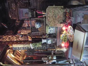 Bức ảnh chụp lại không gian bên trong một ngôi đền hoặc chùa, mang đậm nét cổ kính và trang nghiêm. Trung tâm là một bàn thờ lớn bằng gỗ được chạm khắc công phu. Trên bàn thờ, các vật phẩm thờ cúng được bày biện cẩn thận, bao gồm một nải chuối xanh, một đĩa đựng có thể là tiền vàng mã, và những ngọn nến đang cháy tỏa ánh sáng đỏ ấm áp. Một lư hương lớn cắm đầy nhang (hương). Xung quanh bàn thờ là các vật trang trí khác như một chiếc hộp lớn hình vuông được sơn son thếp vàng, các lọ lộc bình bằng sứ hoa văn xanh trắng, và chân nến bằng đồng. Trên các cấu trúc gỗ như hoành phi và câu đối có khắc nhiều chữ Hán hoặc chữ Nôm. Một bức hoành phi có thể đọc được các chữ '萬古英靈' (Vạn Cổ Anh Linh). Ở phía dưới bức ảnh, có thể thấy hình ảnh phản chiếu của một người đang mặc áo trắng và đội mũ, dường như là một người đến viếng thăm hoặc hành hương đang đứng quan sát.。（日本語訳）。写真は、古風で荘厳な雰囲気が色濃く漂う、ある祠堂または寺院の内部空間を撮影したものである。中央には、精巧な彫刻が施された大きな木製の祭壇がある。祭壇の上には、一房の青いバナナ、冥銭（紙銭）と思われるものが盛られた皿、そして温かい赤い光を放つ燃えているろうそくなど、お供え物が丁寧に並べられている。大きな香炉には線香（香）がたくさん立てられている。祭壇の周りには、金箔で装飾された朱塗りの大きな四角い箱、青と白の模様の磁器製の飾り壺、そして銅製の燭台といった他の装飾品がある。扁額（ホアン・フィ）や対聯（カウ・ドーイ）のような木造物の上には、多くの漢字またはチュノムが刻まれている。ある扁額には「萬古英靈」（Vạn Cổ Anh Linh）という文字が読み取れる。どうやら見学または巡礼に来て、立って見ている人のようだ。