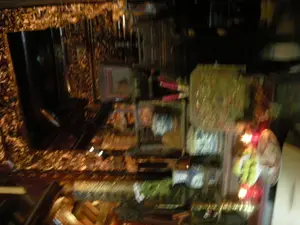 Bức ảnh chụp một cảnh trong nhà, rất mờ và tối, dường như là một bàn thờ. Đây có thể là bàn thờ gia tiên trong một gia đình hoặc bàn thờ tại một nơi thờ cúng công cộng như đền hoặc chùa. Trên bàn thờ có bày biện nhiều vật phẩm. Có thể nhận thấy các bó nhang (hương) với bao bì màu đỏ, một số vật phẩm trông giống hoa quả (có màu xanh và vàng) và các đồ lễ khác được đặt trong giỏ hoặc hộp. Một ánh sáng đỏ rực nổi bật, có thể là từ nến hoặc đèn điện thờ. Cấu trúc xung quanh bàn thờ có các chi tiết trang trí màu vàng, trông giống như được chạm khắc tinh xảo. Mặc dù hình ảnh rất mờ và bị rung, có thể thấy thấp thoáng một vài vật thể giống như bình gốm sứ, trong đó có một chiếc mang hoa văn xanh trắng. Không có chữ viết nào có thể đọc được. Khung cảnh này thể hiện một không gian dành cho hoạt động thờ cúng tâm linh.。（日本語訳）。写真は屋内の光景を写しており、非常にぼやけて暗く、祭壇のようです。これは家庭の祖先の祭壇、または寺院や仏塔のような公共の礼拝所の祭壇である可能性があります。祭壇の上には多くの品物が供えられています。際立つ明るい赤色の光があり、それは蝋燭または祭壇用の電灯からのものかもしれません。祭壇の周りの構造物には黄色の装飾的な細部があり、精巧に彫刻されているように見えます。写真は非常にぼやけて手ブレしていますが、陶磁器の壺のような物体がいくつかかすかに見え、その中には青と白の模様が入ったものも一つあります。読み取れる文字はありません。この光景は、精神的な崇拝活動のための空間を表しています。