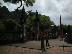 Bối cảnh là một khoảng sân rộng được lát gạch Tàu màu đỏ, nằm trong một khu di tích lịch sử hoặc đền chùa. Lối vào được đánh dấu bởi hai cột đá lớn, được chạm khắc công phu. Trên thân cột có khắc các dòng chữ Hán hoặc Nôm theo chiều dọc nhưng không thể đọc rõ. Bức tường bao quanh cũng được trang trí bằng các hoa văn truyền thống. Trong sân có một vài nhóm du khách đang đi dạo và tham quan. Một người đàn ông mặc áo phông trắng đang đứng cạnh cột đá bên trái. Ở khu vực trung tâm, một nhóm người đang tụ tập, trong đó có một người mặc áo phông màu đỏ và một người đội nón lá. Nhiều lá cờ hội truyền thống, nhiều màu sắc, được cắm trong sân. Phía sau là một công trình kiến trúc có mái ngói và xa hơn là khung cảnh núi non hùng vĩ được cây cối xanh tươi bao phủ.。（日本語訳）。背景は、歴史遺跡地区または寺院の中にある、赤い中国タイルで舗装された広い中庭です。入り口は、精巧に彫刻された2本の大きな石柱で示されています。柱の本体には、縦書きの漢字またはチュノムが刻まれていますが、はっきりと読むことはできません。周囲の壁も伝統的な文様で装飾されています。中庭には、散策したり見学したりしている観光客のグループがいくつかいます。白いTシャツを着た男性が左側の石柱の隣に立っています。中央エリアでは、人々のグループが集まっており、その中には赤いTシャツを着た人とノンラーをかぶった人がいます。中庭には、色とりどりの伝統的な祭りの旗がたくさん立てられています。後方には瓦屋根の建築物があり、さらにその奥には緑豊かな木々に覆われた雄大な山々の風景が広がっています。