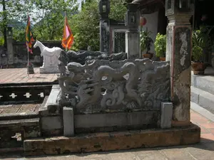 Trên một bức tường ở phía xa bên trái có một biển báo màu xanh với chữ 'WC.' màu trắng. Khung cảnh cho thấy một khoảng sân trong một khu di tích lịch sử hoặc đền, chùa ở Việt Nam. Sân được lát bằng gạch nung màu đỏ. Ở phía trước là một lan can bằng đá được chạm khắc công phu với hình ảnh những con rồng đang uốn lượn trong mây. Hai bên lối đi là các cột trụ bằng đá lớn, đã cũ và nhuốm màu thời gian. Ở phía sau, trên sân, có một bức tượng ngựa trắng (Bạch Mã) bằng đá, có đầy đủ yên cương. Bên cạnh tượng ngựa là hai cột cờ cắm cờ ngũ sắc, loại cờ thường dùng trong các lễ hội truyền thống. Có các bậc tam cấp bằng đá dẫn lên một công trình kiến trúc có mái ngói. Một vài chậu cây cảnh được đặt trên các bậc thềm và một chiếc đèn lồng màu đỏ được treo dưới mái hiên. Không có người trong ảnh, khung cảnh tĩnh lặng và trang nghiêm.。（日本語訳）。左奥の壁には、白い文字で「WC.」と書かれた青い看板があります。この光景は、ベトナムの歴史的遺跡または寺院、仏塔の中庭を示しています。中庭は赤い焼きレンガで敷き詰められています。手前には、雲の中をうねる龍の像が精巧に彫刻された石の手すりがあります。通路の両側には、古びて時の色を帯びた大きな石の柱があります。奥の中庭には、鞍と馬具が完全に備わった、石でできた白馬（バックマー）の像があります。馬の像の隣には、伝統的な祭りでよく使われる種類の五色の旗が掲げられた2本の旗竿があります。瓦屋根の建築物へと続く石の三段の階段があります。いくつかの鉢植えの観葉植物が階段の踊り場に置かれ、一つの赤い提灯が軒下に吊るされています。写真には人がおらず、光景は静かで荘厳です。