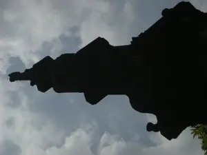 Hình ảnh ghi lại bóng đen của một góc mái nhà thuộc một công trình kiến trúc cổ, được chụp từ góc nhìn thấp hướng lên trời. Phần mái nhà có hình dáng cong vút đặc trưng và các chi tiết trang trí phức tạp, thường thấy trong kiến trúc đền chùa truyền thống của Việt Nam. Toàn bộ công trình kiến trúc chỉ là một khối bóng đen nổi bật trên nền trời xanh với những đám mây trắng xám. Không có bất kỳ văn bản hay chữ viết nào có thể nhận dạng được. Ở góc dưới cùng bên phải, có thể thấy một phần của tán lá cây.。（日本語訳）。画像は、空を見上げる低い視点から撮影された、古い建築物の一角にある屋根の黒い影を記録しています。屋根の部分は、ベトナムの伝統的な寺社建築によく見られる、特徴的に反り上がった形状と複雑な装飾のディテールを持っています。建築物全体は、白灰色の雲がある青空を背景にした、際立った黒い塊にすぎません。認識できるいかなるテキストや文字もありません。右下の一番隅では、樹冠の一部を見ることができます。