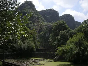 Bức ảnh ghi lại một khung cảnh thiên nhiên yên bình tại một khu vực có núi đá vôi, có thể là ở Ninh Bình. Nổi bật ở hậu cảnh là những dãy núi đá vôi cao, được bao phủ bởi thảm thực vật xanh tươi rậm rạp. Trên sườn núi có thể thấy một công trình trông giống như một cây cầu hoặc đường đi. Bầu trời có mây. Ở tiền cảnh là một hồ nước hoặc ao nhỏ, mặt nước được phủ bởi bèo và lá hoa súng. Xung quanh hồ được xây bờ kè bằng đá, có một cầu thang đá dẫn xuống mặt nước. Cây cối xanh tốt mọc um tùm xung quanh, tạo nên một không gian tĩnh lặng và trong lành. Không có hoạt động của con người và không có chữ viết nào có thể nhận dạng được.。（日本語訳）。写真は、おそらくニンビン省にある石灰岩の山岳地帯の、穏やかな自然の風景を捉えています。背景には、青々とした豊かな植生に覆われた高い石灰岩の山々が際立っています。空は曇っています。前景には湖または小さな池があり、水面は浮き草やスイレンの葉で覆われています。湖の周りは石で護岸が築かれており、水面へと続く石の階段があります。周囲には青々とした木々が生い茂り、静かで清々しい空間を作り出しています。人間の活動は見られず、識別できる文字もありません。