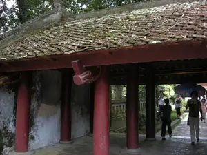 Hình ảnh chụp một hành lang có mái che tại một khu di tích lịch sử, có thể là một ngôi đền hoặc một phần của một công trình kiến trúc cổ ở Ninh Bình. Công trình có kiến trúc cổ đặc trưng với các cột gỗ tròn lớn được sơn màu đỏ. Mái nhà lợp ngói âm dương đã cũ, phủ đầy rêu xanh, lá cây và cành cây khô, cho thấy sự cổ kính và dấu ấn của thời gian. Các bức tường cũng cũ kỹ và loang lổ. Một nhóm người, dường như là khách du lịch, đang đi bộ tham quan dọc theo hành lang lát đá. Một người phụ nữ trong nhóm đang cầm một chiếc ô màu xanh có hoa văn. Phía xa có thể thấy cây cối xanh tươi và một phần lan can bằng đá. Không có chữ viết hay văn bia nào có thể nhận dạng được trong ảnh.。（日本語訳）。歴史的遺跡にある屋根付きの廊下を撮影した写真で、おそらくニンビンにある寺院か、古い建築物の一部であろう。この建造物は、赤く塗られた大きな丸い木製の柱を持つ、特徴的な古い建築様式である。陰陽瓦で葺かれた屋根は古く、青々とした苔、木の葉、枯れ枝で覆われており、古風な趣と時の流れを感じさせる。壁も古びており、まだらになっている。一団の人々、おそらく観光客と思われるが、石畳の廊下に沿って歩いて見学している。グループの中の一人の女性が、模様の入った青い傘を差している。写真の中には、識別できる文字や石碑はない。