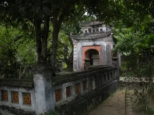 Bức ảnh chụp một công trình kiến trúc cổ, có thể là một ngôi mộ hoặc cổng vào một khu di tích, nằm trong một không gian xanh mát, um tùm cây cối. Công trình chính có cổng vào hình vòm và mái ngói hai tầng đã cũ, nhuốm màu thời gian. Hai bên cổng là hai cột đá có khắc các câu đối bằng chữ Hán, tuy nhiên chữ đã mờ và khó đọc. Có thể nhận ra một vài chữ như '諸將...' và '范...' ở cột bên trái, và các chữ khác ở cột bên phải nhưng không thể xác định rõ. Bên trong cổng có một bệ thờ hoặc bia đá. Bao quanh công trình là một bức tường thấp xây bằng gạch đá, trên tường có các ô cửa sổ trang trí hoa văn bằng đất nung. Khung cảnh tĩnh lặng, không có sự hiện diện của con người hay bất kỳ hoạt động nào.。（日本語訳）。写真は、緑豊かで木々が生い茂る涼しげな空間に佇む、古い建築物を写している。それは墓、あるいは史跡への入り口の門である可能性がある。主な建造物にはアーチ型の入り口の門と、古びて時の色を帯びた二層の瓦屋根がある。門の両脇には漢文の対句が刻まれた二本の石柱があるが、文字は薄れていて読み取りにくい。左側の柱には「諸將...」や「范...」といったいくつかの文字が認識できるが、右側の柱にある他の文字は明確に特定できない。門の内側には祭壇または石碑がある。建造物の周りはレンガと石で造られた低い壁で囲まれており、壁にはテラコッタの文様で装飾された窓枠がある。景色は静寂に包まれ、人の存在やいかなる活動も見られない。