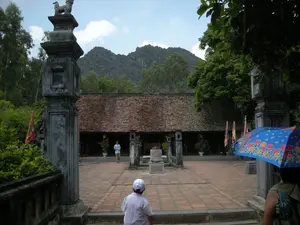 Bức ảnh chụp một khu di tích lịch sử, có thể là một ngôi đền hoặc chùa, với kiến trúc cổ xưa. Ở phía trước là một khoảng sân rộng lát gạch đỏ. Lối vào sân được đánh dấu bằng hai cột đá lớn, trên đỉnh cột bên trái có một bức tượng con Nghê bằng đá. Tòa nhà chính ở phía sau có mái ngói màu nâu đỏ. Có một vài người đang tham quan trong sân: một cậu bé đội mũ trắng đang đi lên bậc thềm, một người đàn ông mặc áo xanh đang đi bộ trong sân, và một người phụ nữ đang che một chiếc ô màu xanh có hoa văn sặc sỡ. Hai bên sân có cắm nhiều lá cờ hội nhiều màu. Khung cảnh xung quanh được bao bọc bởi cây cối xanh tươi và những ngọn núi đá vôi đặc trưng của vùng Ninh Bình ở phía xa. Không có chữ viết nào có thể đọc được trong ảnh.。（日本語訳）。写真は古代建築の史跡を写したもので、寺院か仏塔である可能性があります。手前には赤レンガで舗装された広い中庭があります。中庭への入り口は2本の大きな石柱で示されており、左側の柱の上には石造りのゲの像があります。奥にある本館は赤褐色の瓦屋根です。中庭には数人の観光客がいます。白い帽子をかぶった少年が階段を上っており、青いシャツを着た男性が中庭を歩いており、そして女性が色鮮やかな模様の入った青い傘を差しています。中庭の両側には、色とりどりの祭りの旗が数多く立てられています。写真には読み取れる文字はありません。