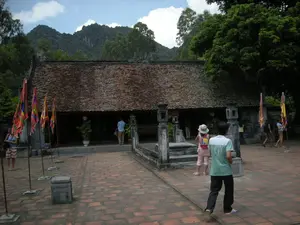 Bối cảnh là một khu di tích lịch sử hoặc đền chùa ở Ninh Bình, với một ngôi nhà cổ kiến trúc truyền thống có mái lợp ngói âm dương màu nâu. Phía trước là một khoảng sân rộng lát gạch đỏ, nơi có nhiều người, dường như là du khách, đang đi tham quan. Trong sân có cắm nhiều lá cờ ngũ sắc, loại cờ thường dùng trong các lễ hội. Một cậu bé mặc áo phông màu xanh lá cây và trắng đang đi bộ trong sân, trong khi những du khách khác, bao gồm một cô bé đội mũ và đeo ba lô, đang đứng gần lối vào có bậc thềm và lan can bằng đá hoặc đi dạo xung quanh. Phía sau công trình là khung cảnh núi đá vôi đặc trưng và cây cối xanh tươi. Không có chữ viết nào có thể đọc được trong hình ảnh.。（日本語訳）。背景はニンビン省にある歴史遺跡または寺院で、そこには茶色の陰陽瓦で葺かれた屋根を持つ伝統的な建築様式の古い家があります。前方には赤いレンガが敷き詰められた広い中庭があり、そこでは観光客と思われる多くの人々が見学しています。中庭には、祭りでよく使われる五色旗が多く立てられています。緑と白のTシャツを着た少年が中庭を歩いている一方で、帽子をかぶりバックパックを背負った少女を含む他の観光客たちは、石の階段と手すりのある入り口の近くに立っていたり、あたりを散策したりしています。建物の後ろには、特徴的な石灰岩の山々と青々とした木々の風景が広がっています。画像内には読み取れる文字はありません。