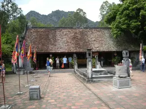 Bối cảnh là một khu di tích lịch sử hoặc đền chùa ở Việt Nam, có thể là ở khu vực Ninh Bình, với kiến trúc truyền thống. Phía trước là một khoảng sân rộng lát gạch đỏ. Một ngôi nhà cổ có mái ngói âm dương màu nâu sẫm và các cột gỗ. Nhiều người, có vẻ là khách du lịch, đang tham quan khu di tích. Một người đàn ông nước ngoài mặc áo xanh đang đi bộ trong sân, tay cầm một chiếc nón lá. Trong sân có cắm nhiều lá cờ ngũ sắc dùng trong lễ hội. Ở phía trước có một lư hương lớn bằng đá, bên trong có nhiều chân hương. Phía sau là cây cối xanh tươi và những dãy núi đá vôi. Không có chữ viết nào có thể đọc được trong ảnh.。（日本語訳）。背景はベトナムの歴史遺跡または寺院で、おそらくニンビン地方にあり、伝統的な建築様式です。前方には、赤いレンガが敷き詰められた広い中庭があります。暗褐色の陰陽瓦の屋根と木製の柱を持つ古い家があります。多くの人々、おそらく観光客が、遺跡を観光しています。青いシャツを着た外国人男性が、ノンラーを手に持ち、中庭を歩いています。中庭には、祭りで使われる多くの五色の旗が立てられています。前方には大きな石造りの香炉があり、その中には多くの線香の燃え残りがあります。後方には青々とした木々と石灰岩の山々があります。写真の中には読み取れる文字はありません。