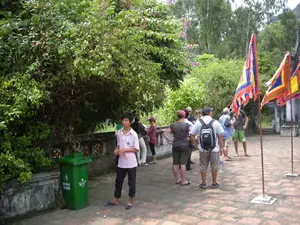 Trên thùng rác màu xanh lá cây có dòng chữ: 'HÃY BỎ RÁC VÀO THÙNG' và logo 'MTĐT'. Bối cảnh là một khoảng sân lát gạch tại một địa điểm tham quan, có thể là một ngôi chùa hoặc di tích lịch sử ở khu vực Ninh Bình. Xung quanh có nhiều cây cối xanh tươi và một giàn hoa giấy. Một nhóm du khách, có vẻ bao gồm cả người nước ngoài, đang tụ tập tham quan. Họ mặc trang phục thoải mái như áo phông, quần short và một người đàn ông đeo ba lô. Ở phía bên phải, có một số lá cờ hội (cờ ngũ sắc) được cắm trên cột. Đứng tách biệt với nhóm, một cậu bé người Việt Nam đang đứng gần thùng rác.。（日本語訳）。緑色のゴミ箱の上には、「ゴミはゴミ箱に入れましょう」という文字と「MTĐT」というロゴがある。背景は、ニンビン地方にある観光地のタイル張りの庭で、おそらく寺院か史跡である。周りには青々とした木々がたくさんあり、ブーゲンビリアの棚が一つある。彼らはTシャツ、ショートパンツといった楽な服装をしており、一人の男性はバックパックを背負っている。右側には、何本かの祭りの旗（五色旗）が柱に立てられている。集団から離れて、一人のベトナム人の少年がゴミ箱の近くに立っている。