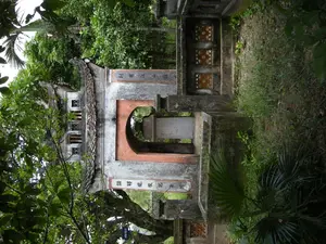 Đây là hình ảnh một công trình kiến trúc cổ, có khả năng là cổng vào của một khu lăng mộ hoặc khu tưởng niệm. Công trình được xây bằng đá và xi măng, mang dáng vẻ cổ kính. Cấu trúc chính bao gồm một lối đi hình vòm ở trung tâm, hai bên là hai cột trụ lớn. Trên hai cột trụ có khắc các dòng chữ Hán Nôm theo chiều dọc. Cột bên phải (nhìn từ ngoài vào) có khắc chữ '諸將隨駕歿於陣' (Chư tướng tuỳ giá một ư trận), có nghĩa là 'Các tướng đi theo vua đã chết tại trận'. Cột bên trái khắc chữ '别有表碑記其事' (Biệt hữu biểu bi ký kỳ sự), nghĩa là 'Có bia riêng ghi lại sự việc đó'. Phía sau cổng vòm có thể thấy một tấm bia đá (bia ký). Mái cổng được lợp ngói ống truyền thống với các góc mái cong vút. Xung quanh công trình là cây cối um tùm, bao gồm cả cây cọ và nhiều loại cây lá rộng khác. Nền đất mọc đầy cỏ dại, cho thấy sự cổ kính của di tích. Không có người hay hoạt động nào diễn ra trong ảnh.。（日本語訳）。これは古い建築物の画像で、おそらく陵墓または記念区域の入り口の門でしょう。この建造物は石とセメントで建てられており、古風な趣を呈しています。主な構造は、中央のアーチ型の通路と、その両側にある2本の大きな柱で構成されています。2本の柱には、縦書きで漢字・チュノムの文字列が刻まれています。（外から見て）右側の柱には「諸將隨駕歿於陣」（Chư tướng tuỳ giá một ư trận）と刻まれており、その意味は「王に従った諸将は戦場で亡くなった」です。左側の柱には「别有表碑記其事」（Biệt hữu biểu bi ký kỳ sự）と刻まれており、その意味は「その出来事を記録した別の石碑がある」です。アーチ門の後ろには、石碑（碑文）が見えます。門の屋根は伝統的な筒瓦で葺かれており、屋根の角は反り上がっています。建造物の周りには、ヤシの木や他の多くの広葉樹を含む木々が生い茂っています。地面には雑草が生い茂っており、遺跡の古さを示しています。写真の中には人や活動は見られません。