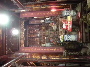 Bên trong một ngôi đền hoặc miếu thờ có kiến trúc truyền thống. Toàn bộ không gian được làm chủ yếu bằng gỗ sẫm màu, được chạm khắc tinh xảo. Ở trung tâm là một bàn thờ lớn, nhiều tầng, được trang trí công phu. Trên bàn thờ có các vật phẩm thờ cúng, bao gồm một ống sứ men lam trắng đựng đầy trái cây (có vẻ là táo xanh), một ống sứ tương tự cắm một cây nến đỏ đang cháy, và một số bánh kẹo được bày trên đĩa. Phía trên bàn thờ có treo các bức hoành phi sơn son thếp vàng. Một bức có ghi các ký tự Hán Nôm: '丁皇上廟 黎皇下祠' (Đinh Hoàng Thượng miếu, Lê Hoàng Hạ từ), gợi ý đây là đền thờ Vua Đinh và Vua Lê. Các bức hoành phi khác có các dòng chữ như '正氣參天' (Chính khí tham thiên) và '萬古流芳' (Vạn cổ lưu phương). Phía sau bàn thờ là các bức vách bằng gỗ được chạm trổ phức tạp. Ở phía trước, có thể thấy các loại binh khí nghi lễ có cán dài. Khung cảnh tĩnh lặng, thể hiện một không gian thờ tự trang nghiêm, được chiếu sáng bởi đèn điện hiện đại.。（日本語訳）。伝統的な建築様式の祠堂または廟の内部。空間全体は主に暗い色の木材で作られ、精巧な彫刻が施されている。中央には、精巧に装飾された大きな多層の祭壇がある。一つの扁額には「丁皇上廟 黎皇下祠」（ディン・ホアン・トゥオン廟、レ・ホアン・ハ祠）という漢字（チュノム）が書かれており、これがディン王とレ王を祀る祠堂であることを示唆している。他の扁額には「正氣參天」（せいきさんてん）や「萬古流芳」（ばんこりゅうほう）のような文字がある。祭壇の後ろには、複雑な彫刻が施された木製の壁がある。現代の電灯に照らされた、荘厳な祭祀空間を示す静かな光景である。