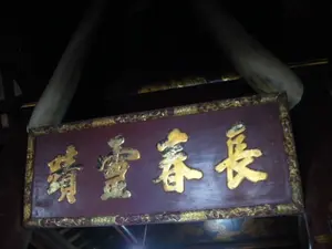 Bức ảnh chụp cận cảnh một bức hoành phi bằng gỗ được treo trong một không gian tối, có khả năng là bên trong một ngôi đền, miếu hoặc đình. Bức hoành phi có nền màu đỏ sẫm, khung viền được chạm khắc và mạ vàng. Trên đó có khắc nổi bốn chữ Hán lớn được thếp vàng, đọc từ phải sang trái là: "Trường Xuân Linh Từ" (長春靈祠), có nghĩa là "Đền thiêng Trường Xuân". Phía trên bức hoành phi có treo một cặp ngà voi lớn.。（日本語訳）。扁額は暗赤色の背景で、縁には彫刻が施され金メッキが塗られています。その上には金箔を施した四つの大きな漢字が浮き彫りにされており、右から左へ「Trường Xuân Linh Từ」（長春靈祠）と読み、意味は「長春の聖なる祠」です。扁額の上方には、一対の大きな象牙が掛けられています。