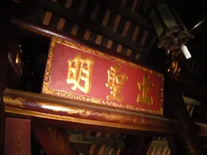 Bên trong một công trình kiến trúc cổ, có thể là đền hoặc chùa, với kết cấu chủ yếu bằng gỗ sẫm màu. Ở vị trí trung tâm phía trên là một bức hoành phi lớn sơn son thếp vàng. Trên nền đỏ của bức hoành phi có ba chữ Hán lớn được mạ vàng: 明聖主 (Minh Thánh Chủ). Viền của bức hoành phi được trang trí bằng các họa tiết hoa văn mạ vàng tinh xảo. Phía trên bên phải, một chiếc đèn lồng kiểu cổ được treo trên trần nhà, bên trong có lắp một bóng đèn huỳnh quang compact hiện đại. Không có hoạt động nào của con người được ghi lại, khung cảnh tĩnh lặng, mang tính trang nghiêm.。（日本語訳）。古い建築物、おそらく祠か寺の内部で、主に濃い色の木材で構成されている。上部中央には、朱塗り金箔仕上げの大きな扁額がある。扁額の赤い背景の上には、金メッキが施された3つの大きな漢字「明聖主」（ミン・タイン・チュー）がある。扁額の縁は、精巧な金メッキの模様で装飾されている。右上には、古風な提灯が天井から吊るされており、その中には現代的な電球形蛍光灯が取り付けられている。人間の活動は記録されておらず、光景は静かで荘厳である。