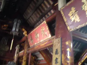 Bên trong một công trình kiến trúc cổ, có thể là đền, chùa hoặc đình làng. Toàn bộ kết cấu được làm bằng gỗ, với các cột, xà và mái nhà có màu nâu sẫm. Các chi tiết kiến trúc như cột và cổng được sơn son thếp vàng, trang trí bằng các hoa văn màu vàng. Có nhiều biển hiệu bằng gỗ được treo lên, bao gồm các bức hoành phi (biển ngang) và câu đối (biển dọc). Các biển này có nền màu đỏ hoặc màu gỗ sẫm, trên đó có khắc các chữ Hán Nôm mạ vàng. Do chất lượng hình ảnh bị mờ và góc chụp, các ký tự này không thể đọc rõ. Không có người hay hoạt động nào được ghi lại trong ảnh.。（日本語訳）。古い建築物の内部、それは寺院、仏塔、あるいは村の集会所かもしれません。全ての構造は木でできており、柱、梁、屋根は濃い茶色です。柱や門などの建築の細部は、朱漆と金箔で塗られ、黄色の模様で装飾されています。多くの木製の看板が掲げられており、それには横額（横長の扁額）と対聯（縦長の扁額）が含まれます。これらの看板は赤色または濃い木色の背景で、その上には金メッキが施された漢字・チュノムが刻まれています。画像の品質がぼやけていることと撮影角度のため、これらの文字ははっきりと読み取れません。写真には人や活動は記録されていません。